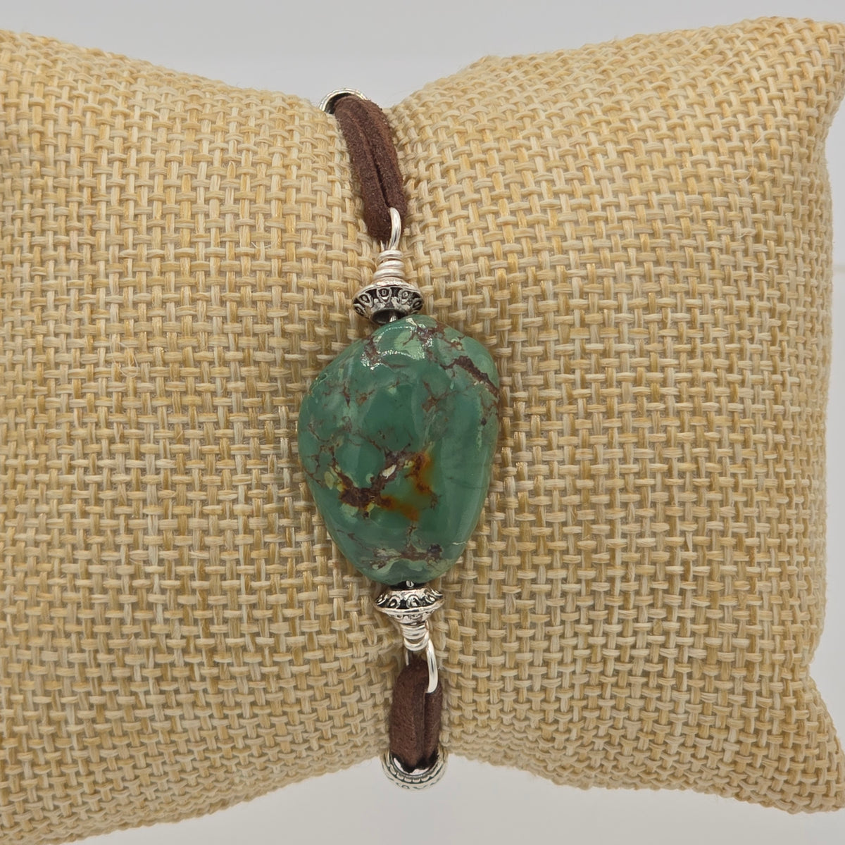 A green marbled stone bead bracelet with brown leather cord and silver-toned metal spacers, displayed on a beige woven cushion.